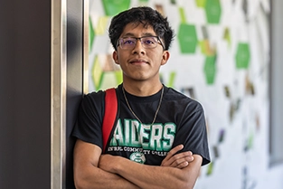 Male student wearing a Central Community College Raiders shirt, leaning against the wall with his arms crossed