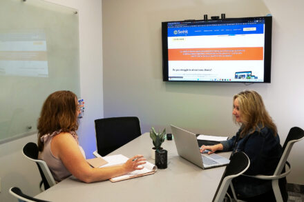 Two people working in small office with one laptop, and wall-mounted monitor