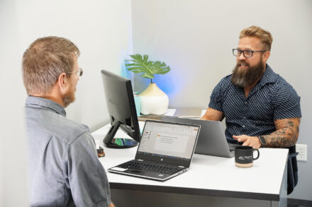 Two men sitting at a table, talking while working on their laptops