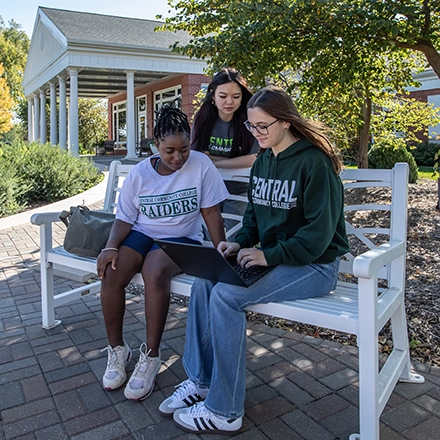 Two girls sitting on a bench looking at a laptop and a third girl standing behind the bench.