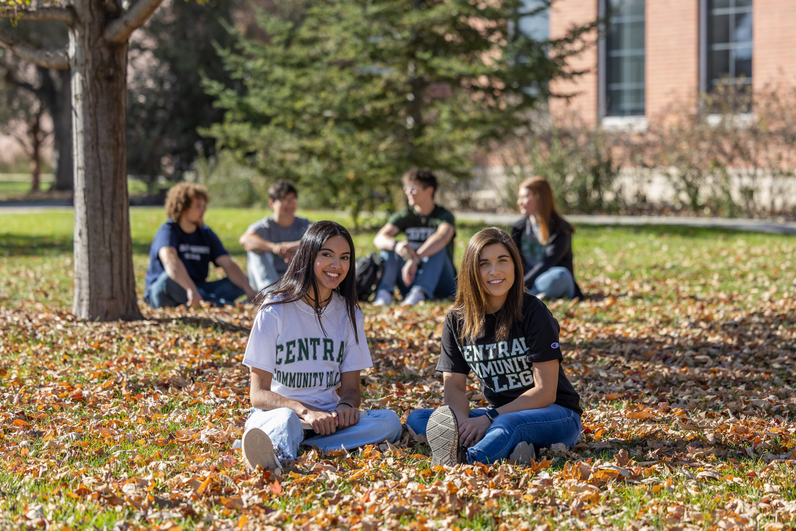 Two students sit on the grass on campus during the fall leaves.