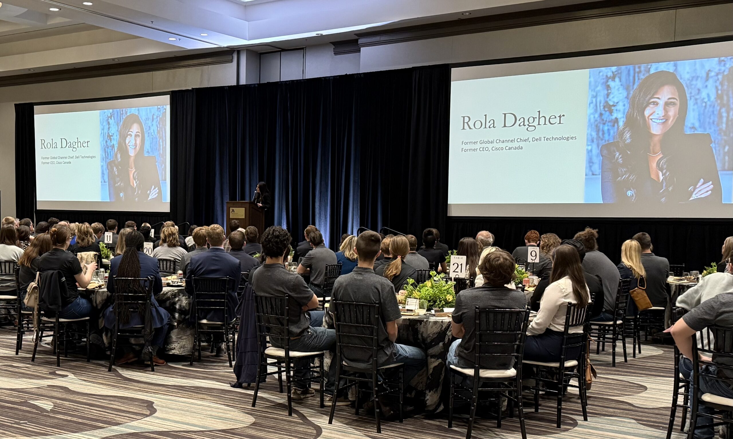 Speaker Rola Dagher presents on stage as audience sits at round tables in a conference hall.