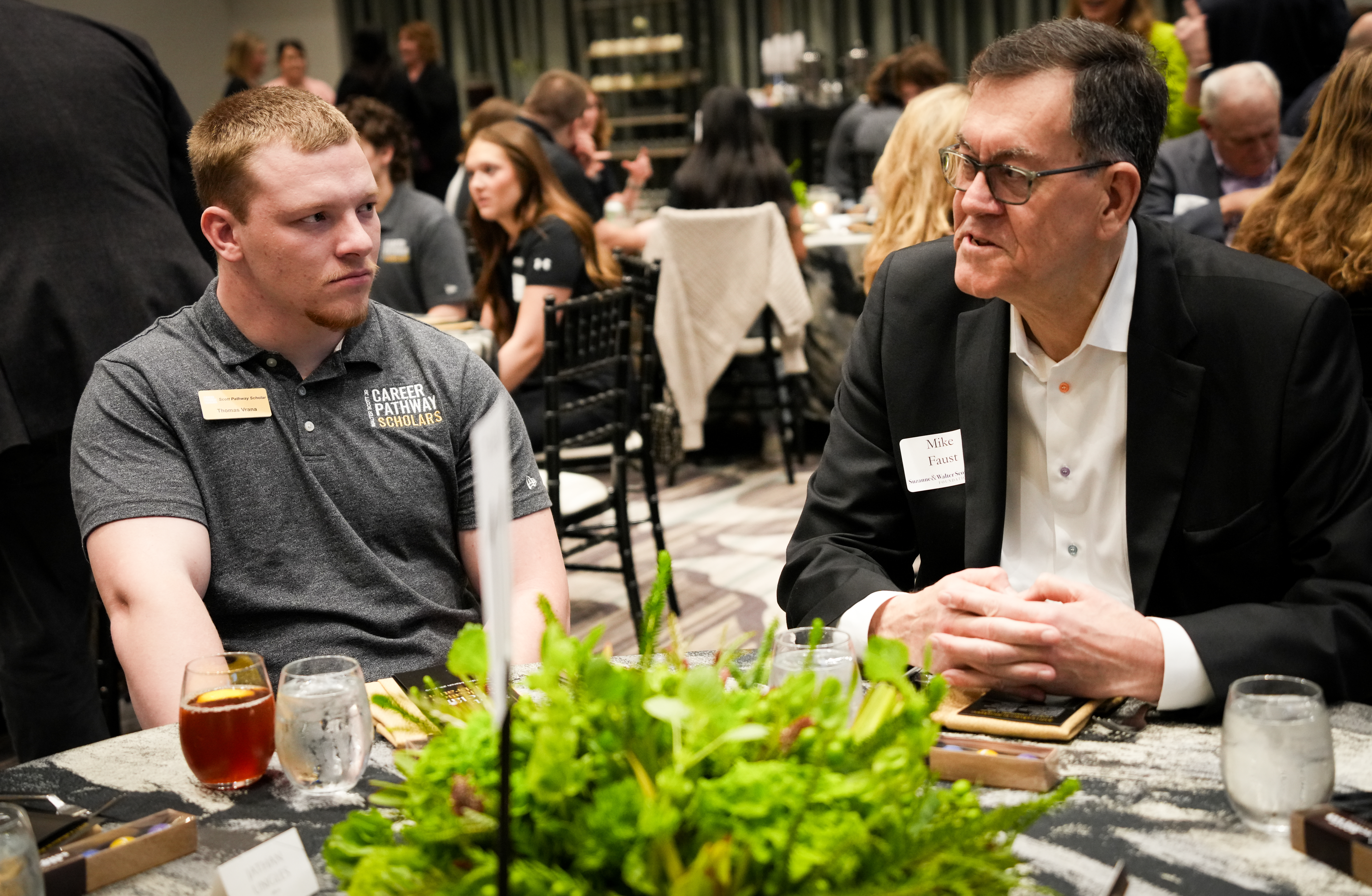 Two people talk across a table with greenery centerpiece at a large banquet event.