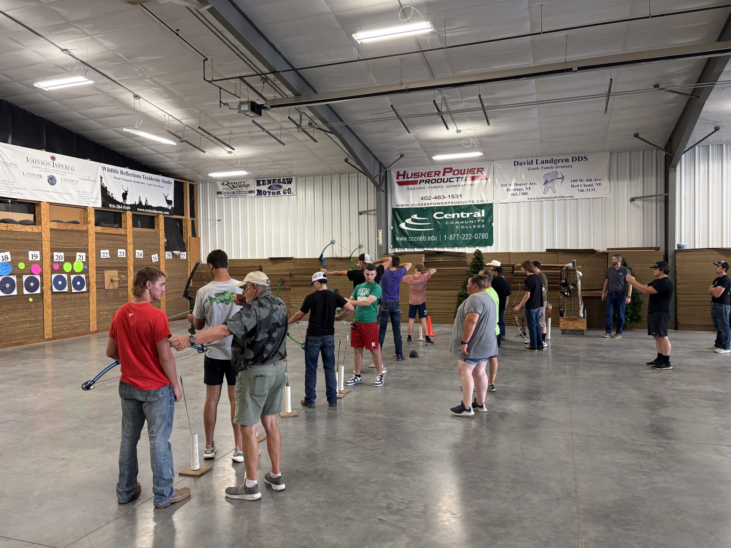 Archery participants stand in a large indoor range aiming bows toward targets on the far wall.