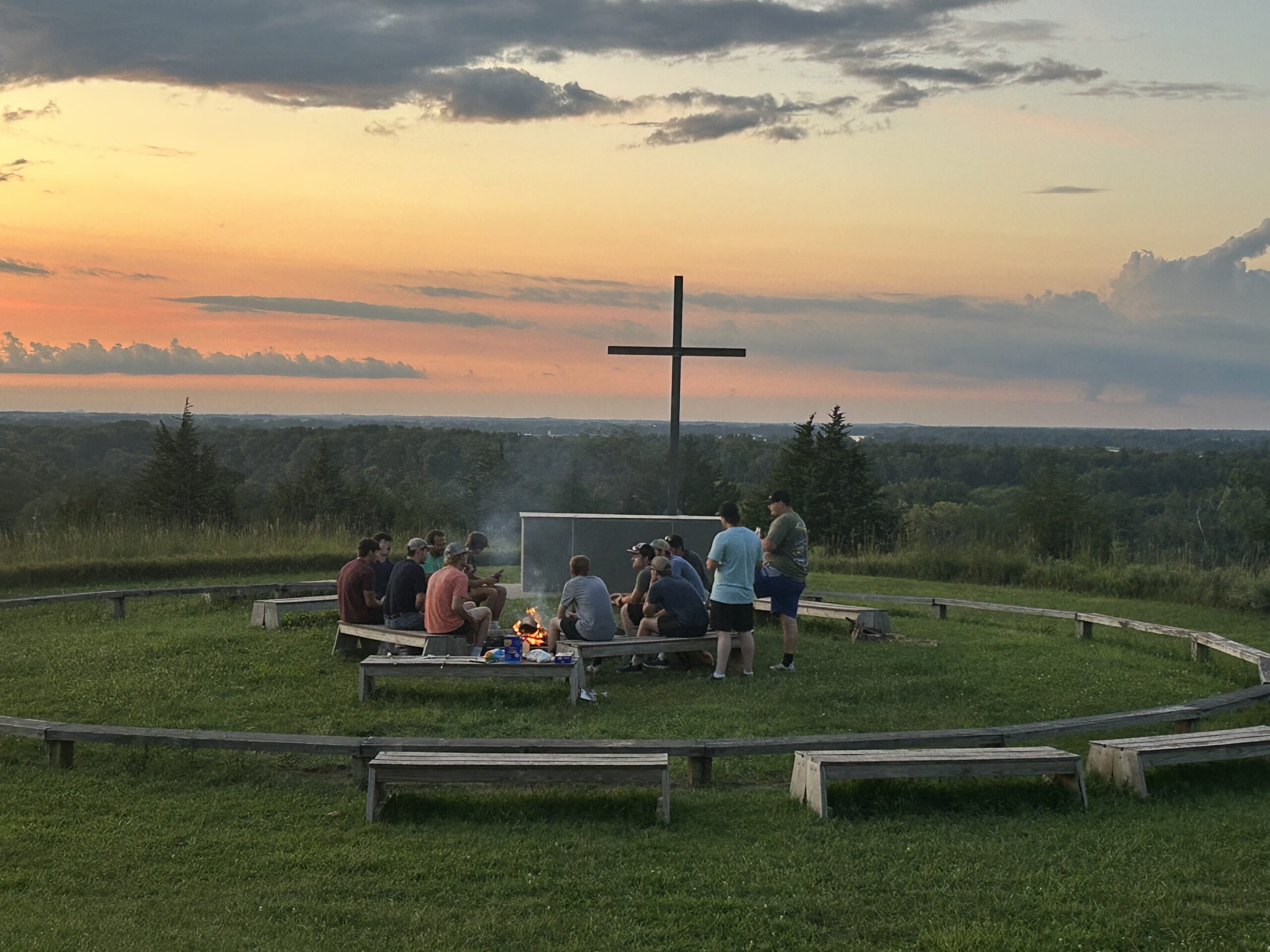 Group sits around a campfire near a large cross as the sun sets.