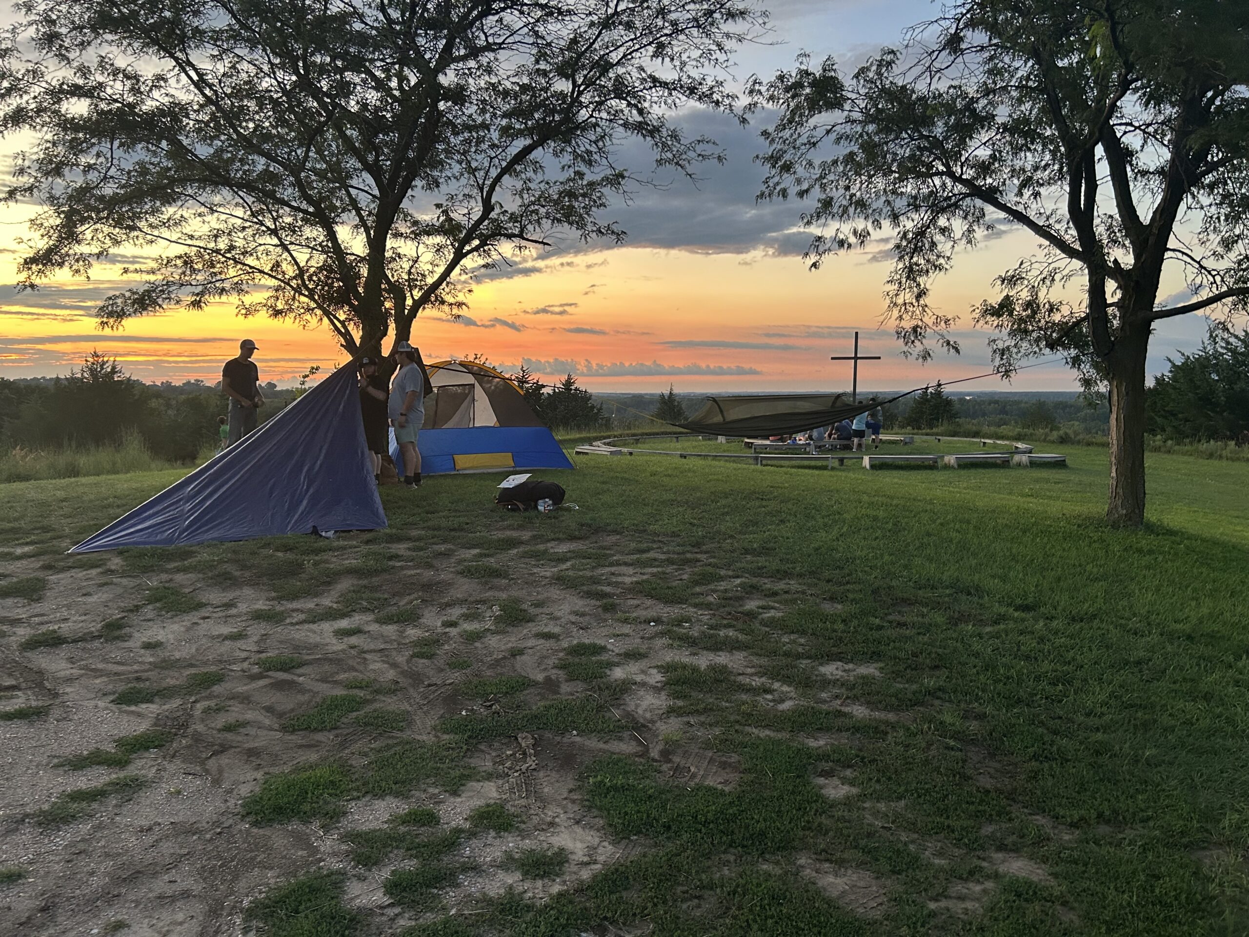 Tents set up on a grassy hilltop campsite at sunset with a cross in the distance.