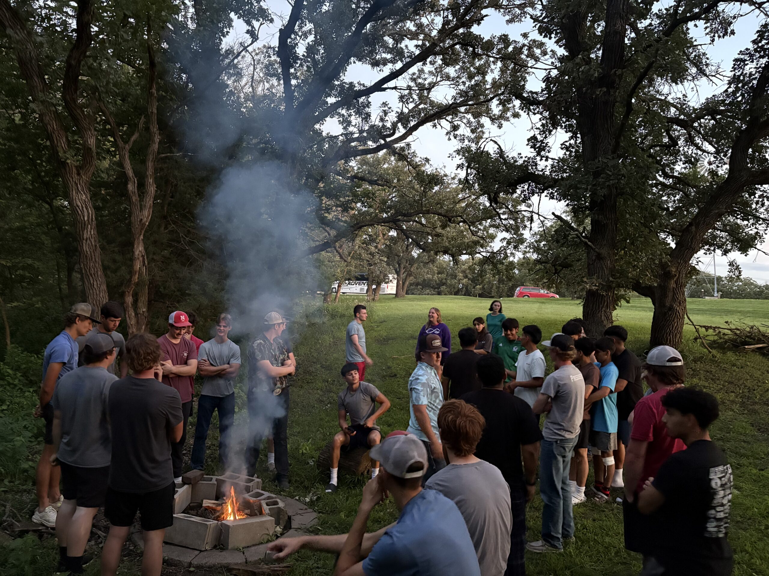 Group gathered around a campfire outdoors, smoke rising among trees in a grassy area