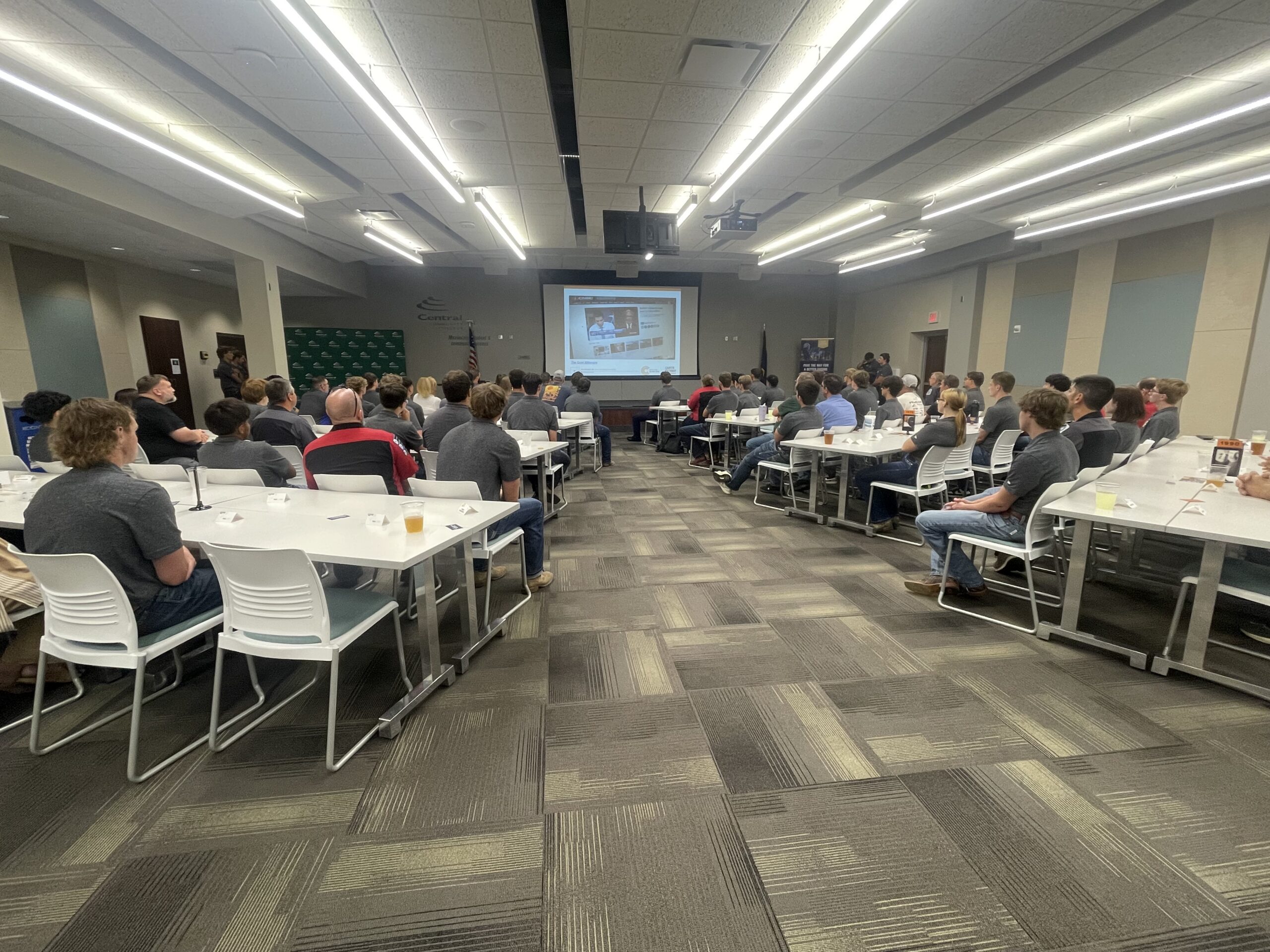 Wide classroom with rows of tables and seated attendees facing a projector screen.