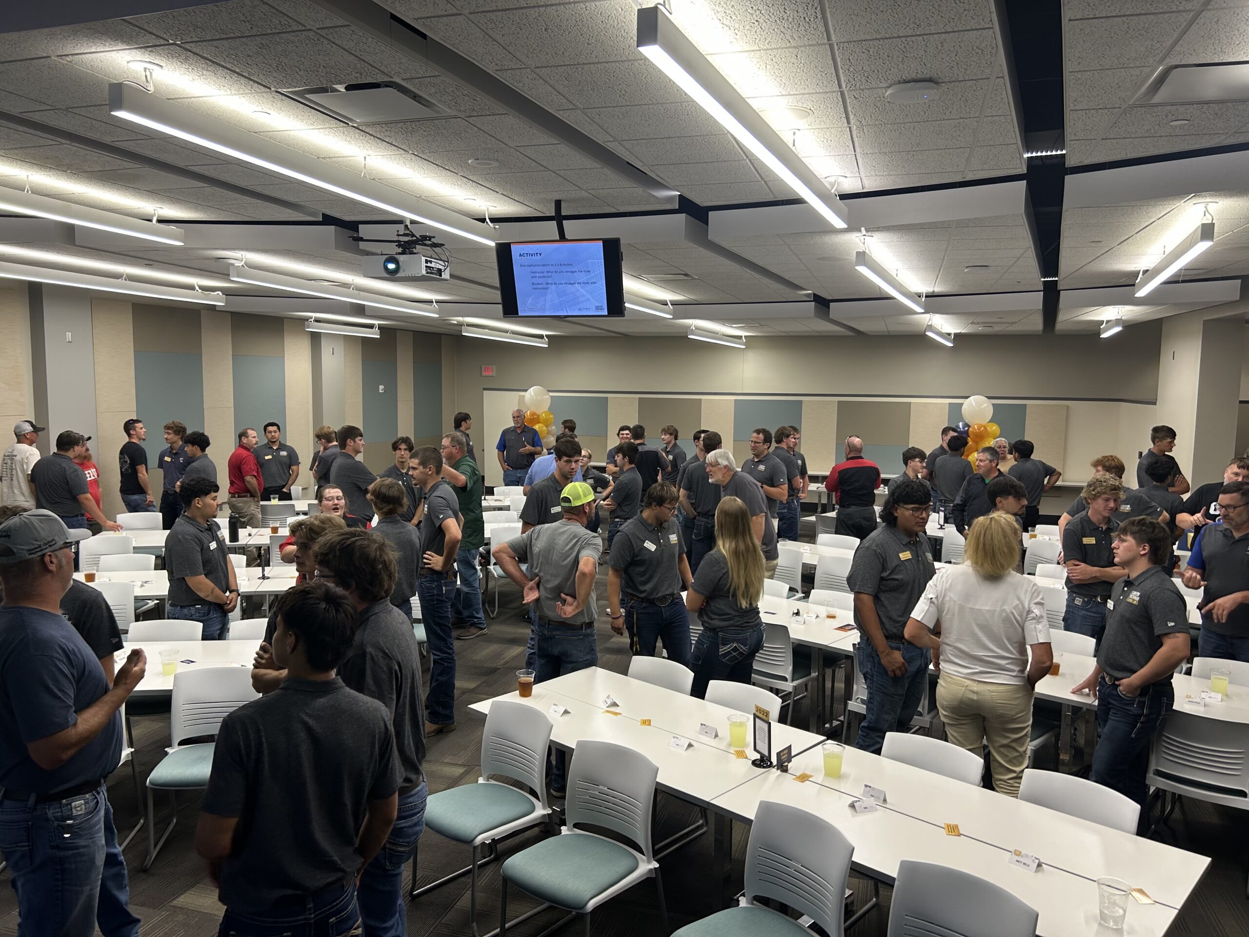 Crowded classroom with groups standing around tables, open laptops, and screens at front.