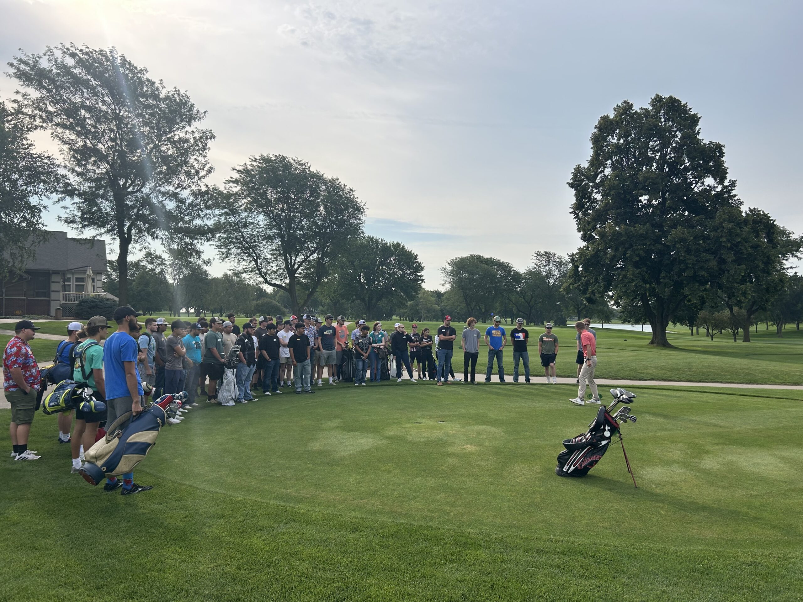 Group of students gathered on a golf course listening to an instructor demonstrating a swing.
