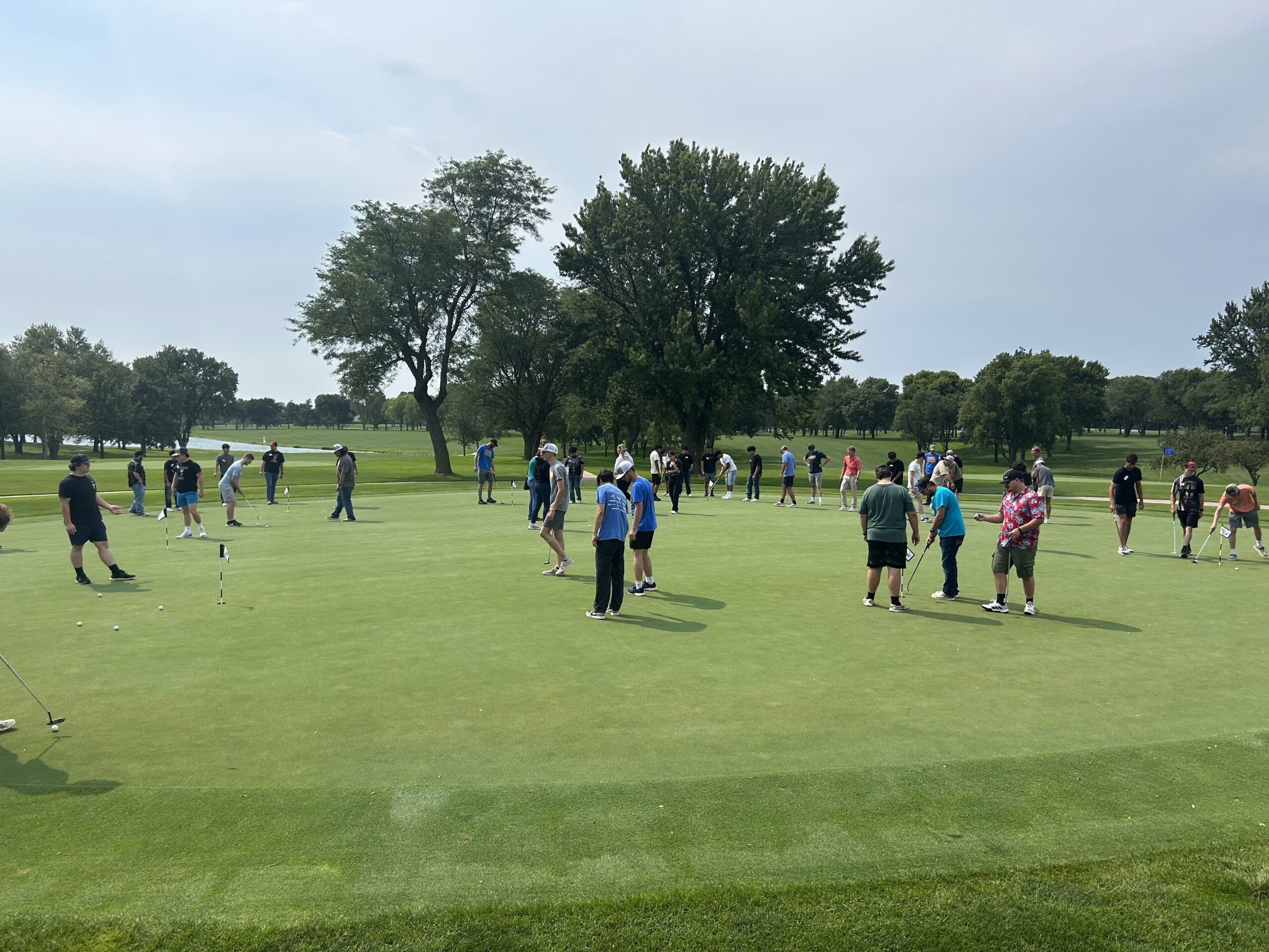 Group of people gathered on a putting green during a golf event, trees lining the course.