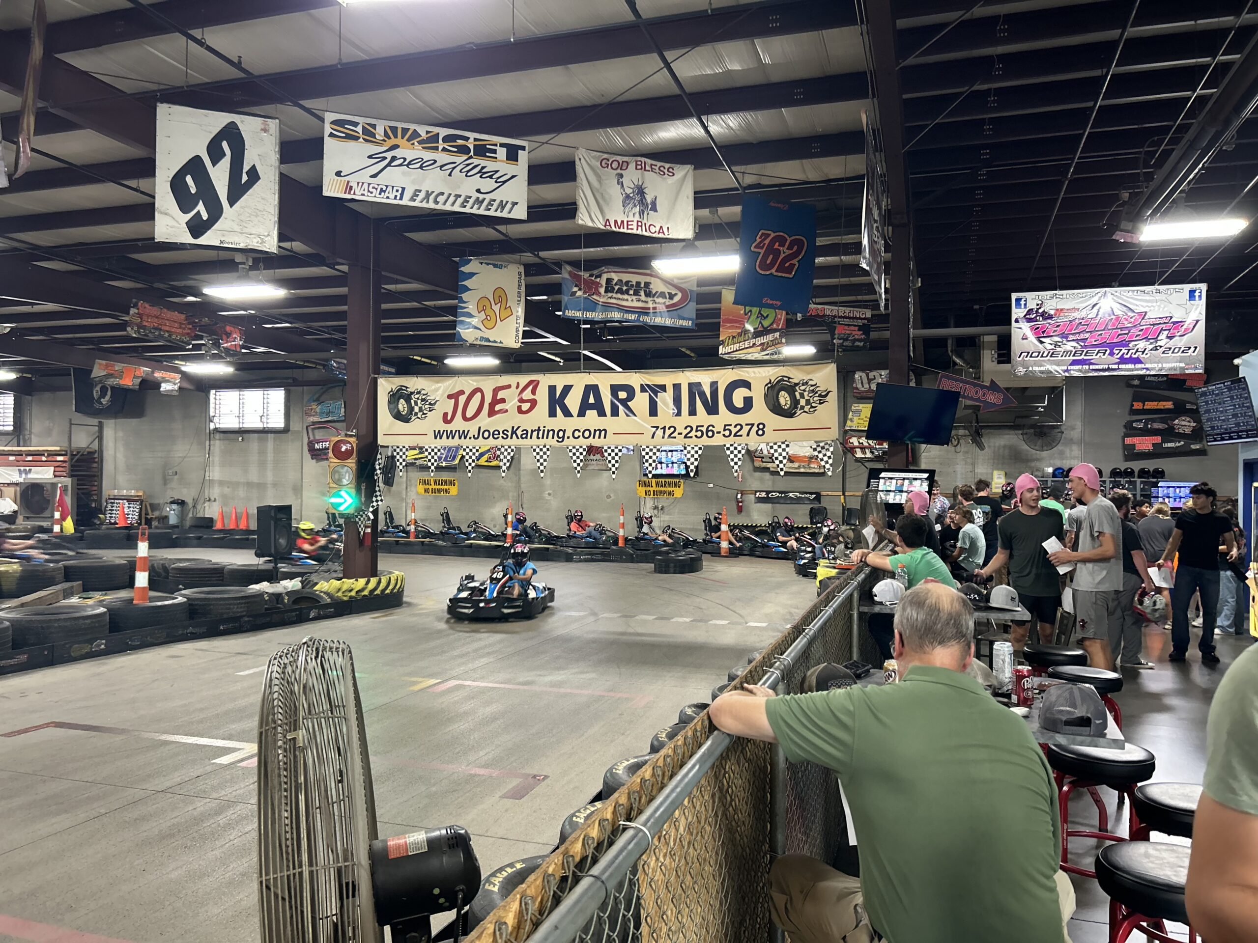 Indoor go-kart track with racers on the course and spectators behind a barrier at Joe's Karting