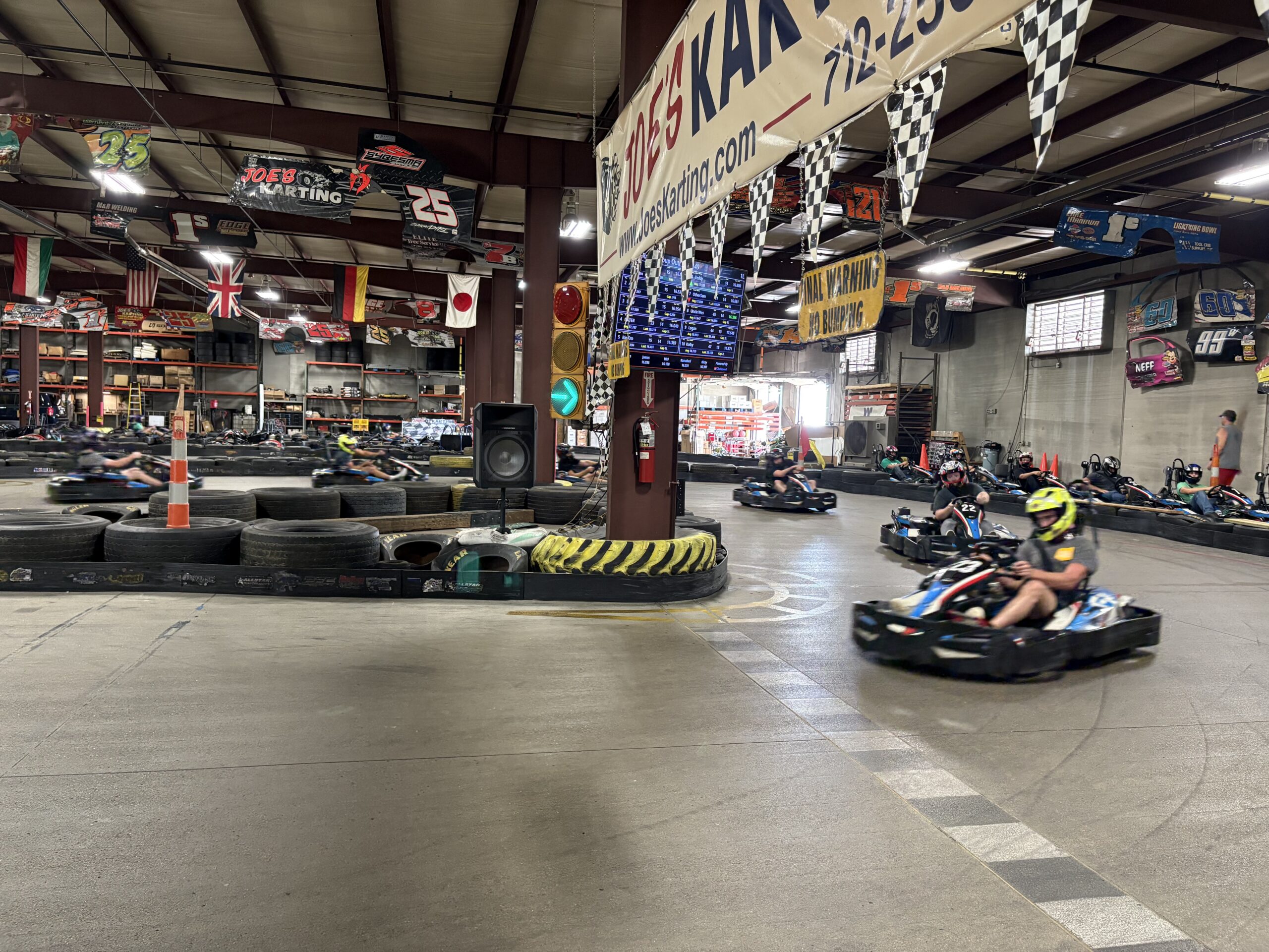 Indoor go-kart racers speed around a track with checkered banners overhead.