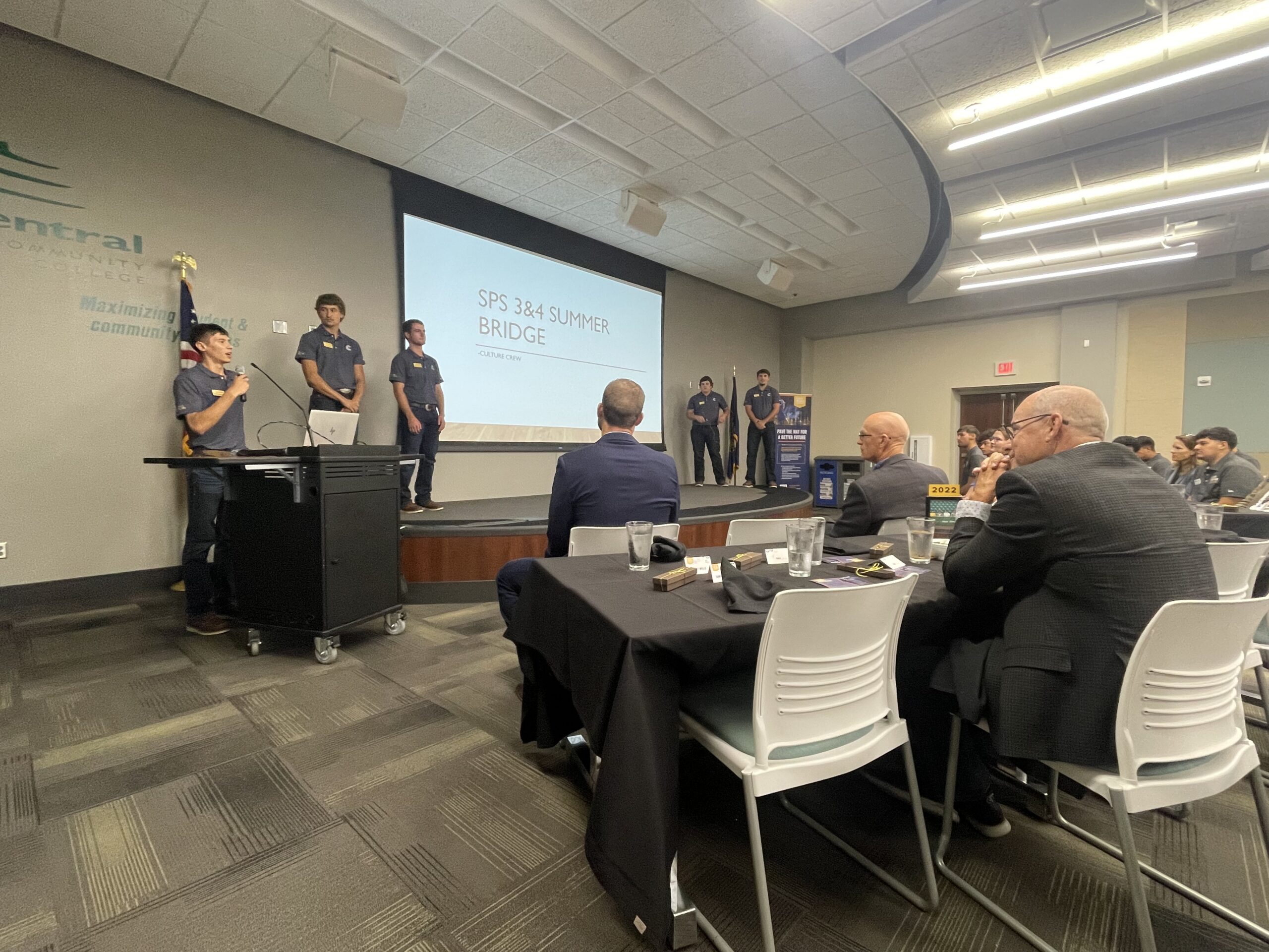 Students present on stage to an audience during a summer bridge kickoff event.