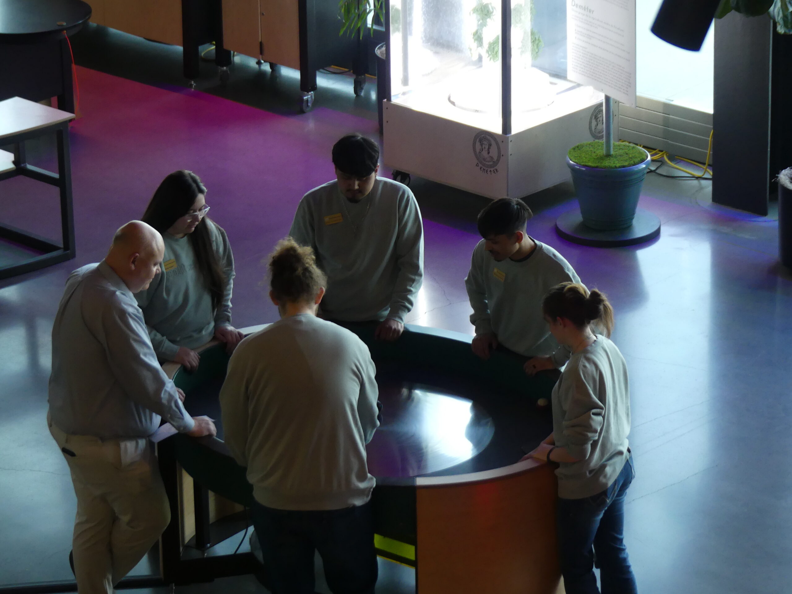 Six people stand around a round interactive table exhibit in a museum space.