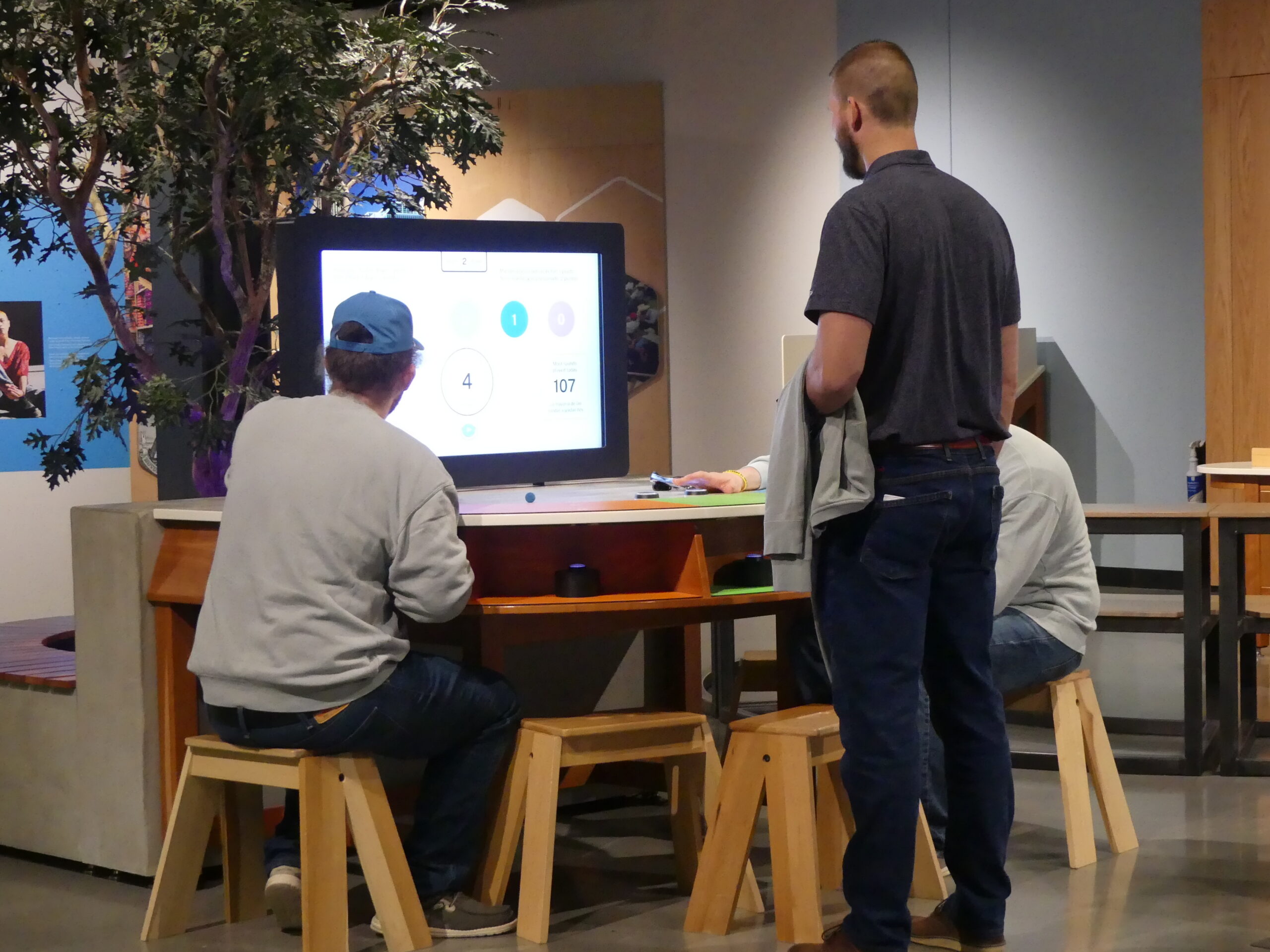 People gather at a digital tabletop display while one person stands nearby observing.