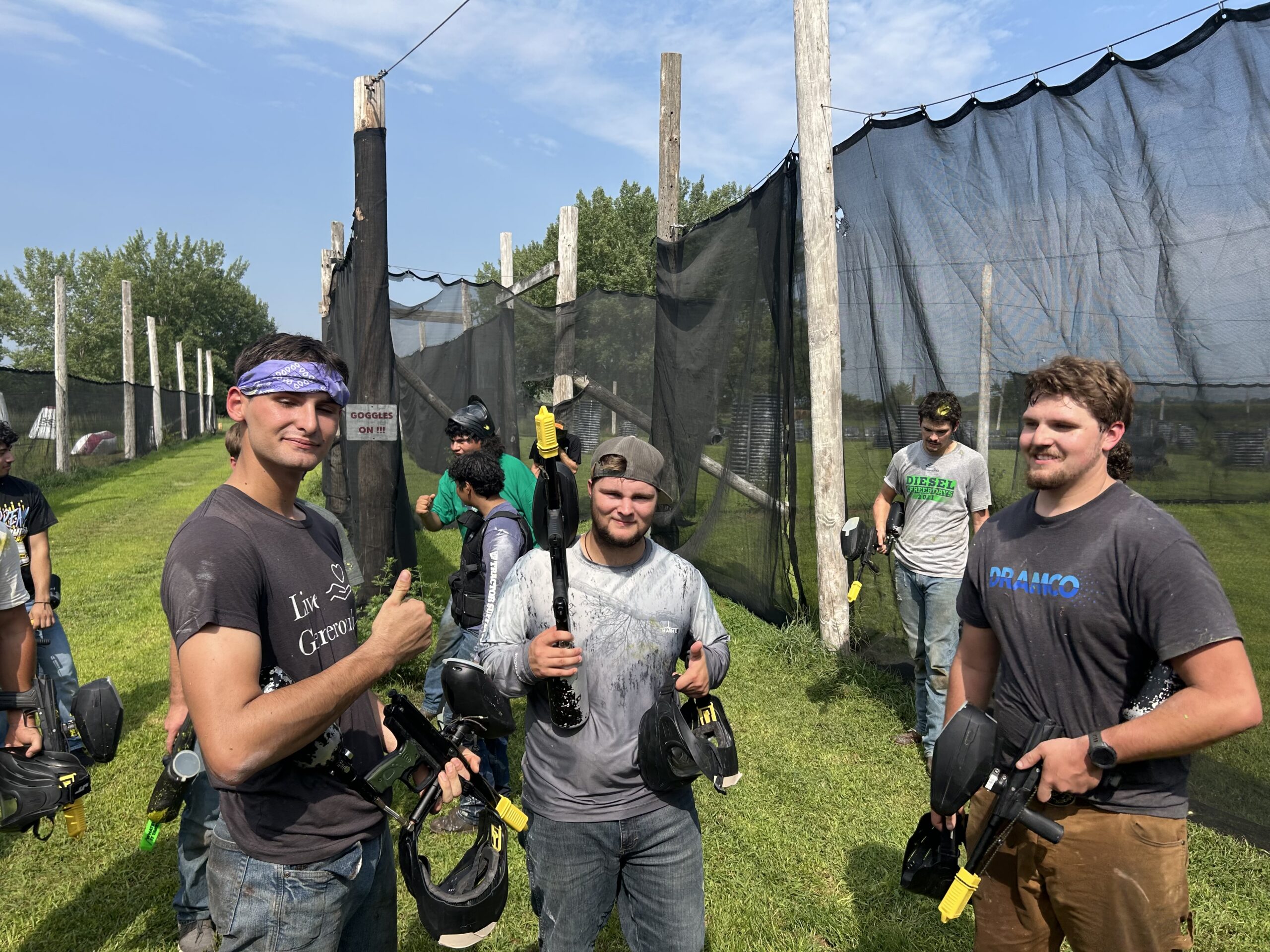 Group of people holding paintball markers standing on a grassy field near netted barriers.