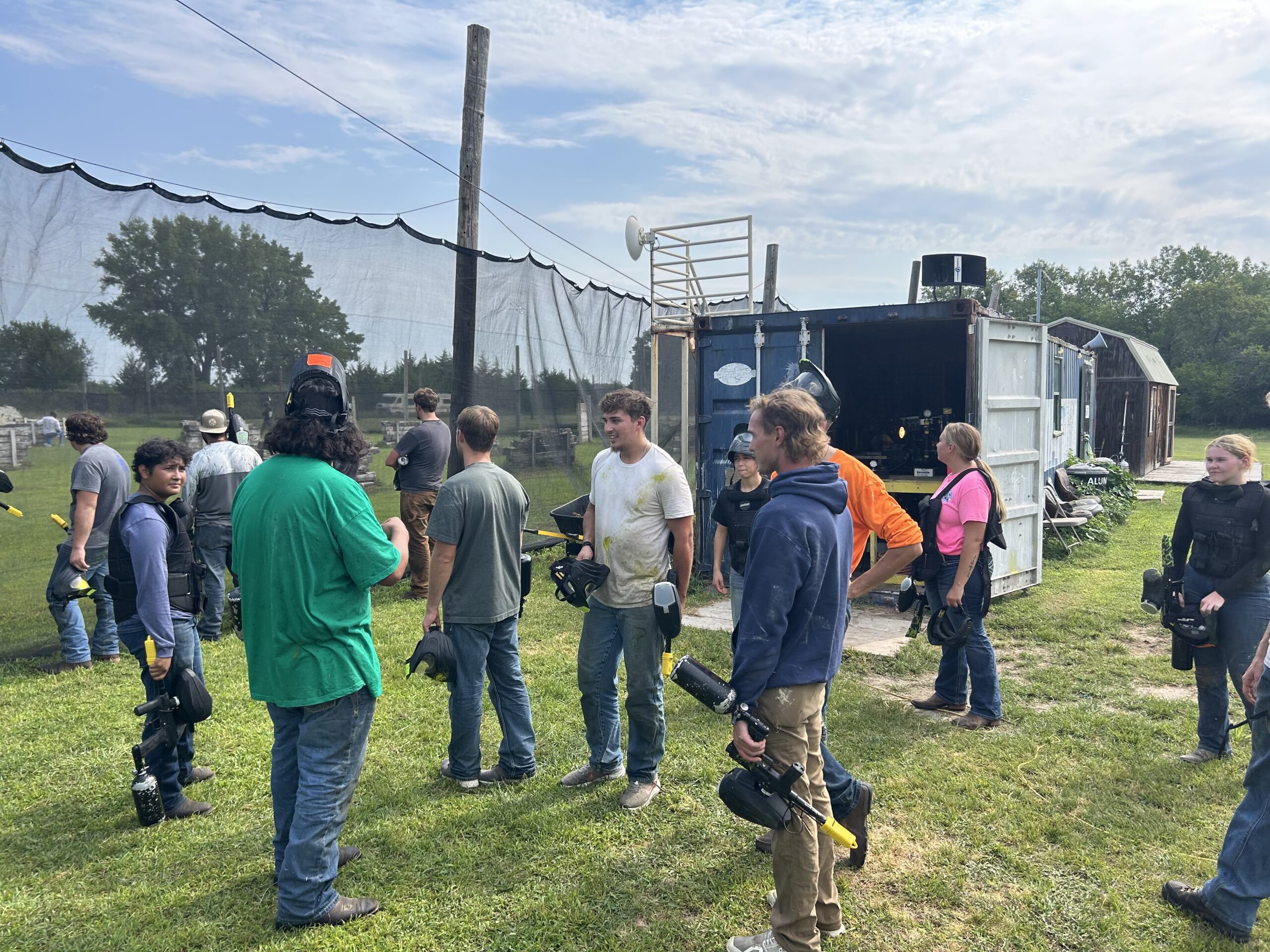Participants hold paintball gear while gathering near a field and equipment trailer.