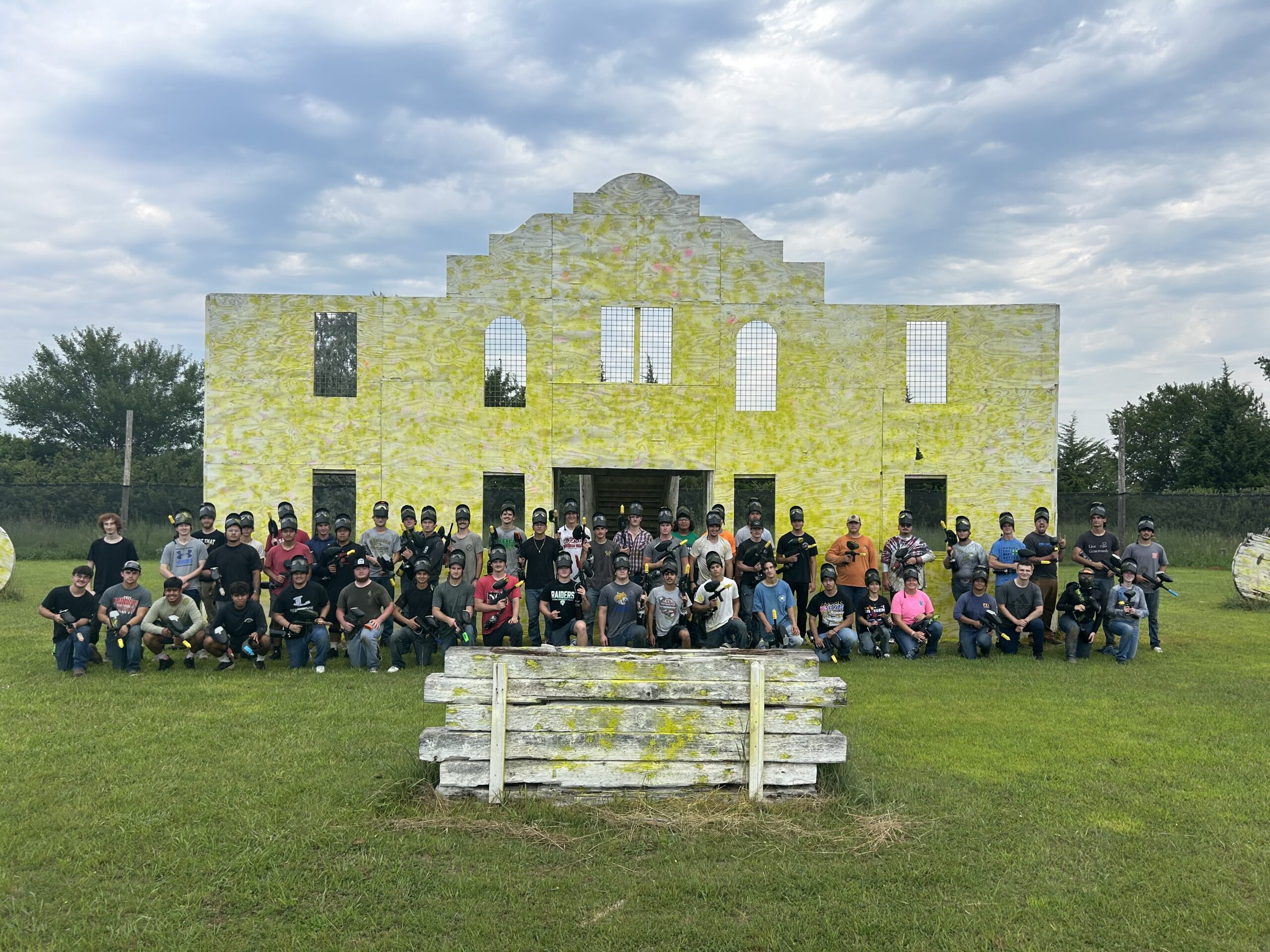 Large group of people posing on grass in front of a yellow paintball field structure under a cloudy sky
