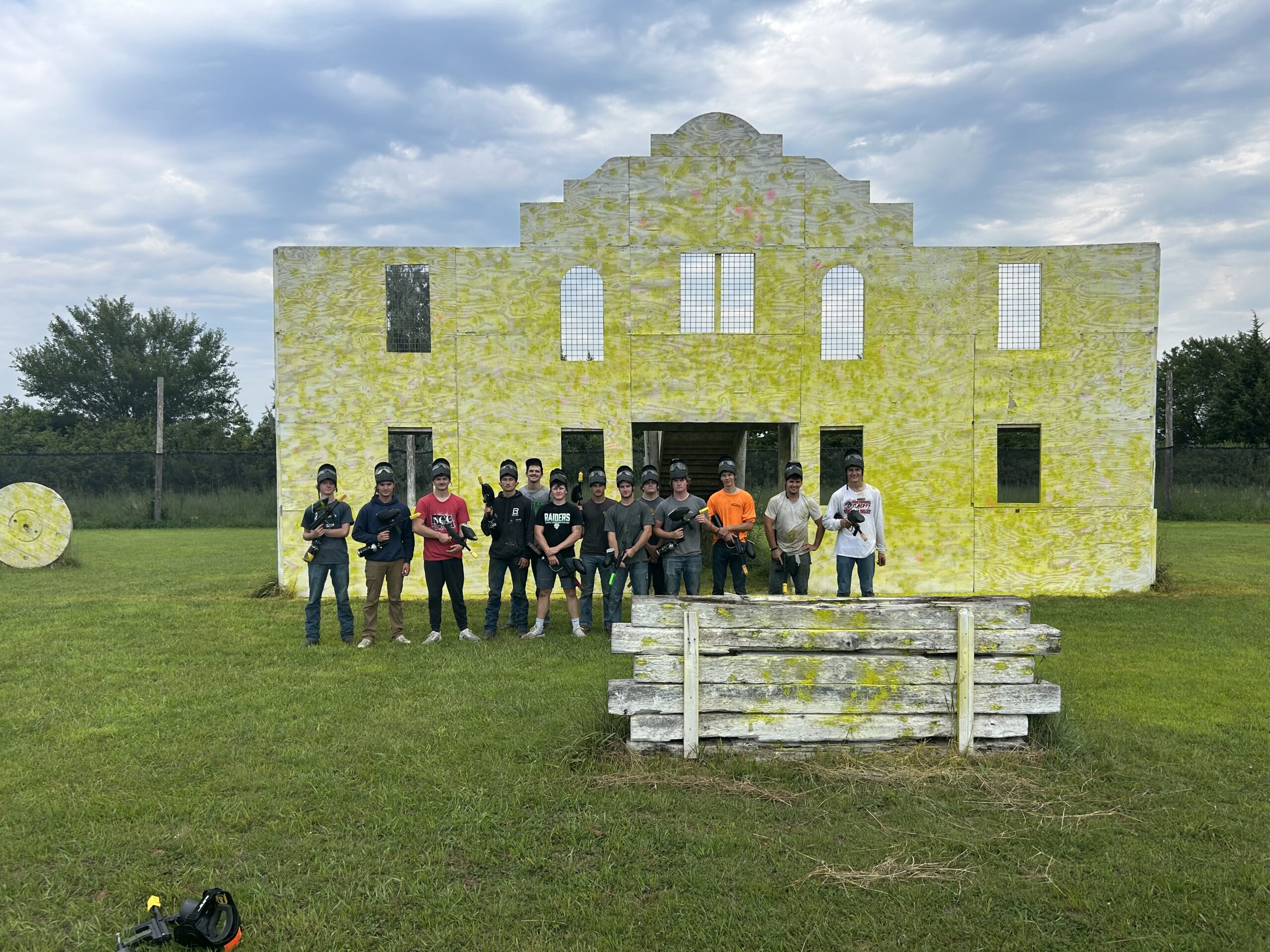 Group poses in front of a paint-splattered target building at an outdoor paintball field.