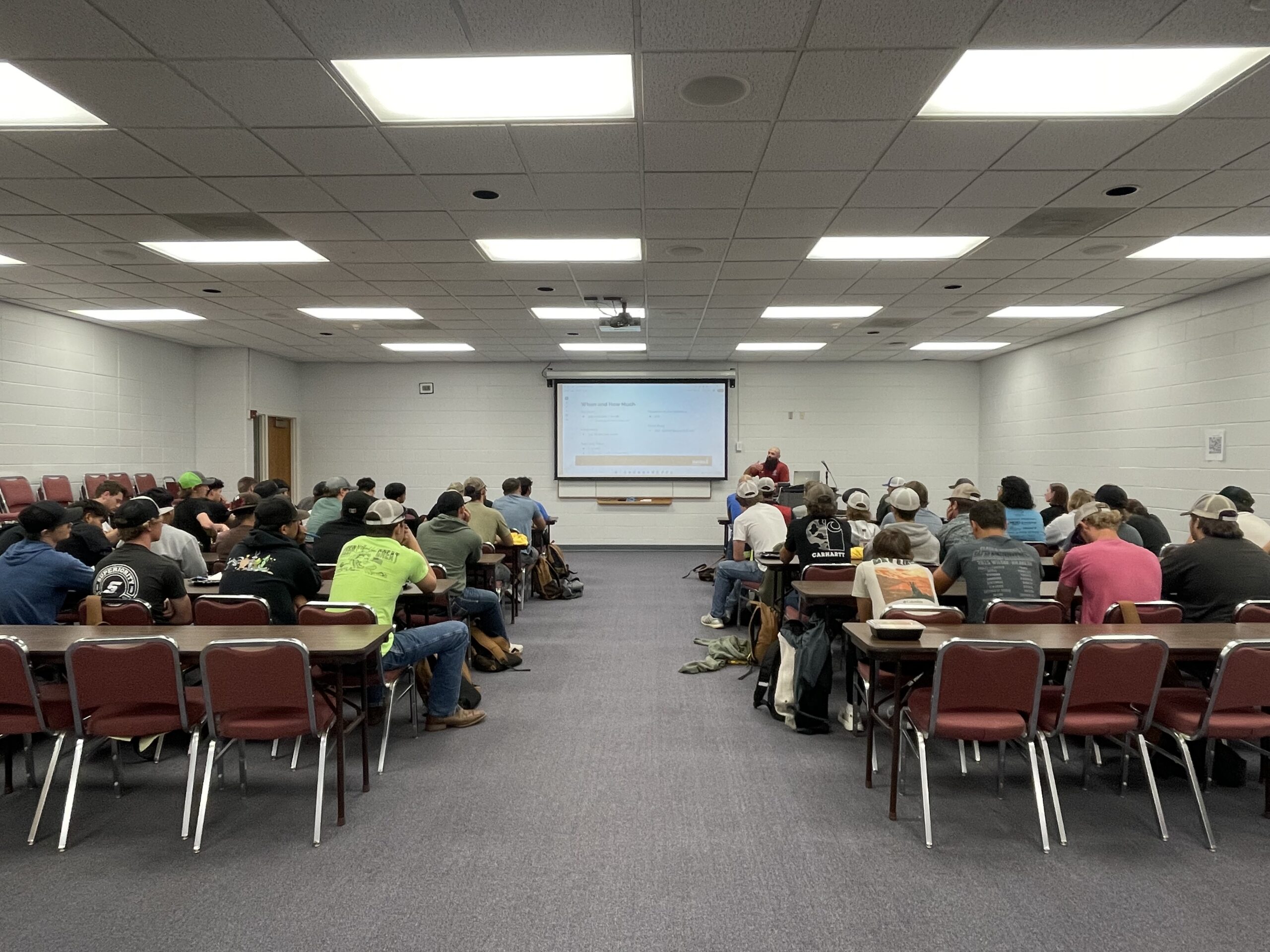 Attendees seated at long tables in a large room, watching a presentation on a projector.