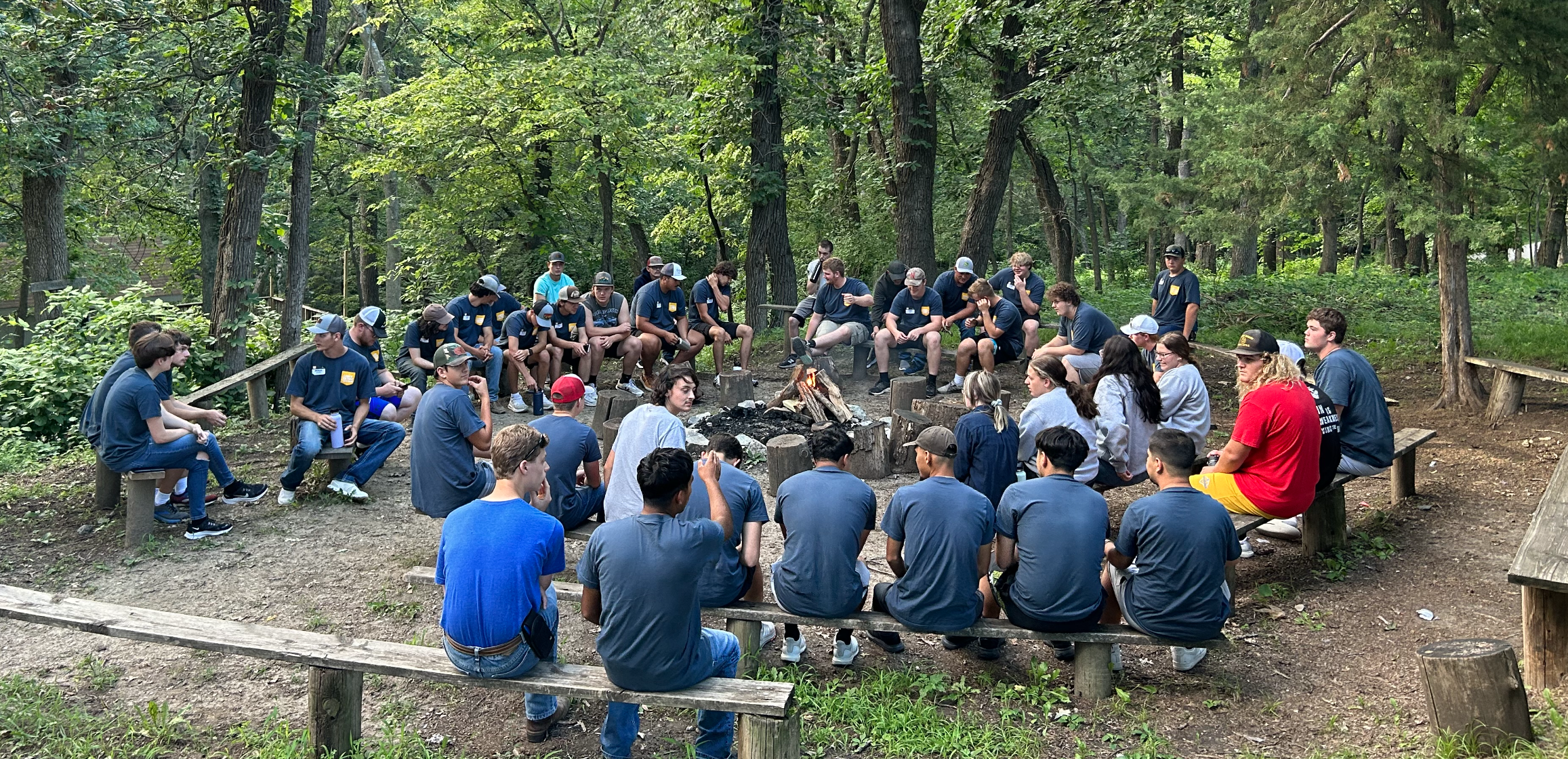 Group sits in a circle outdoors, gathered around a campfire area in the woods.
