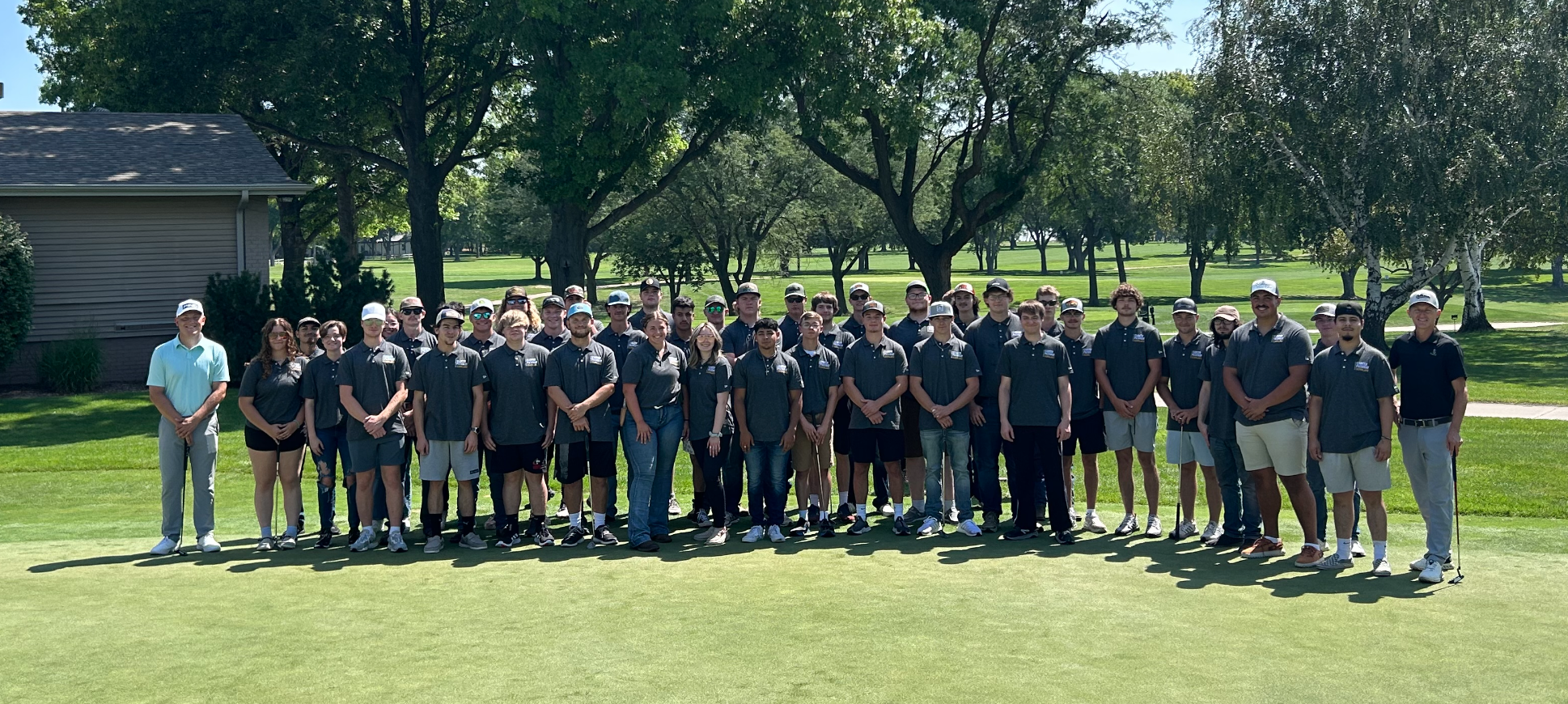 Large group poses for a photo on a golf course under trees and clear sky.