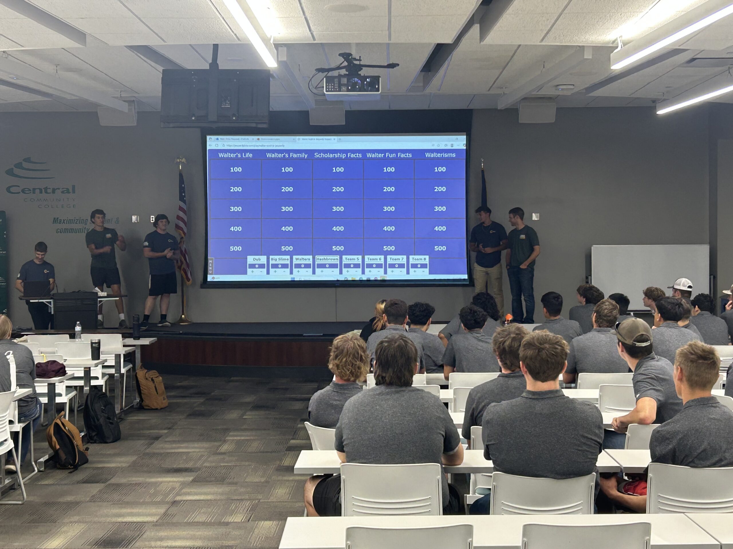 Audience seated in classroom watching presenters beside a large screen showing a data table.